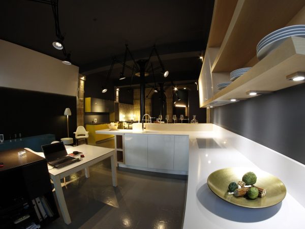 Local con cocina blanca abierta, puesto de trabajo y techo negro con luminarias industriales