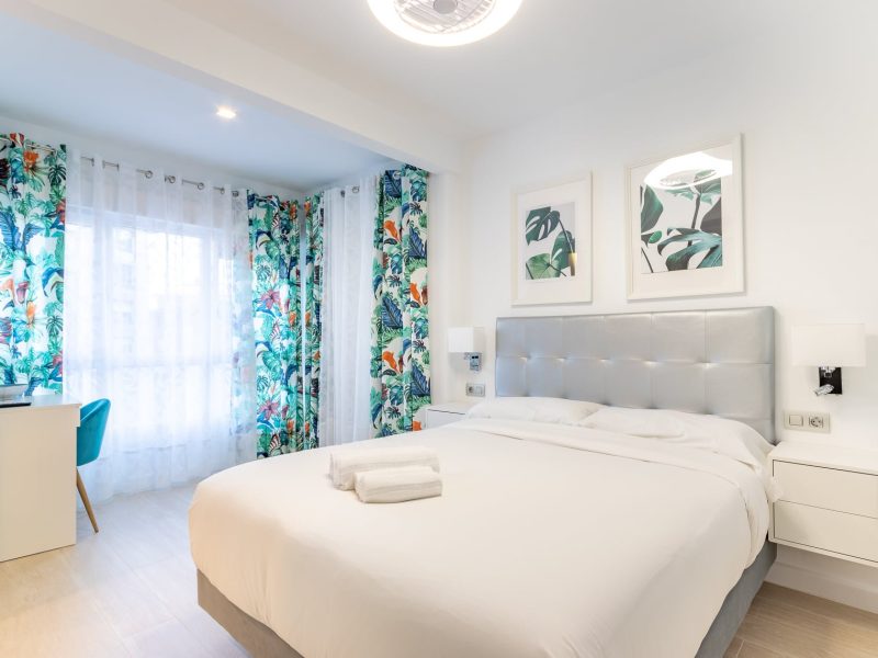 Dormitorio de vivienda turística con cama blanca, cabecero tapizado y cortinas de estampado tropical