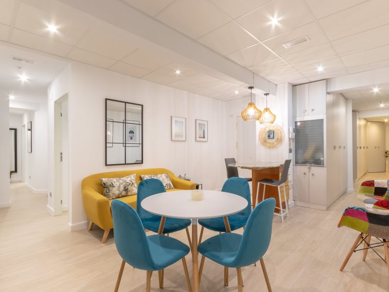 Salón comedor de vivienda turística con sillas turquesa, sofá mostaza y mesa redonda blanca